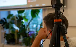 A webcam on a tripod in the foreground; behind it, a person watches the projected screen.